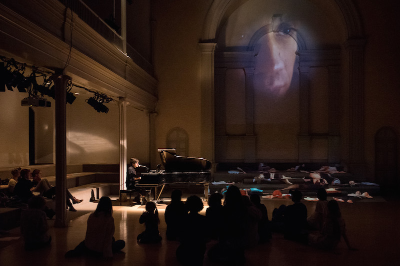 Morgan Bassichis plays on the piano. Audience members sit on the floor. A projection of a man's side profile is projected on the back wall